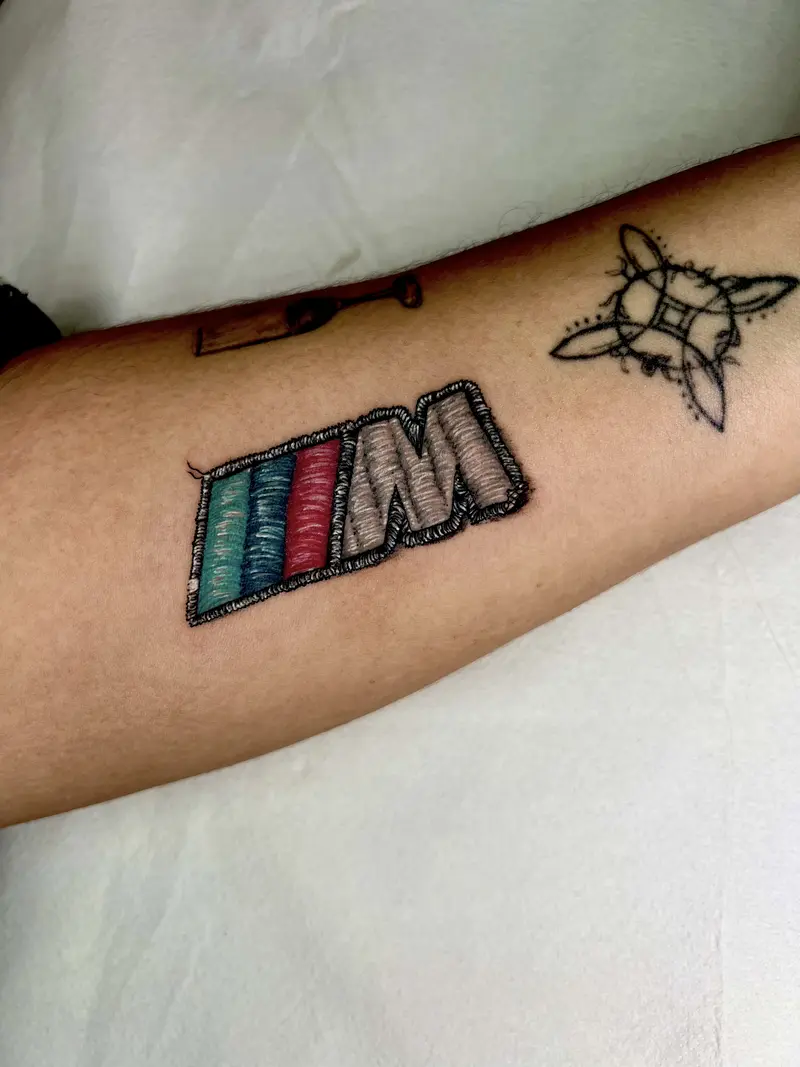 Tatuaje estilo parche bordado del logotipo “M” con franjas tricolor, efecto hilo y relieve.